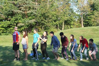 Radcliffe Choral Society and Harvard Glee Club members standing in a single line playing games on a grassy lawn during choir retreat. 