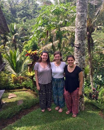 Three Radcliffe Choral Society members standing together in front of a large jungle in Indonesia. All three singers are wearing different iterations of the same outfit. 