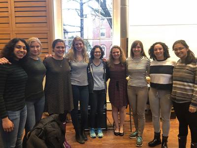 Nine Radcliffe Choral Society members standing in a semi-circle, posing for a picture in front of a window and whiteboard in Harvard's Holden Chapel. All nine people are wearing striped shirts.