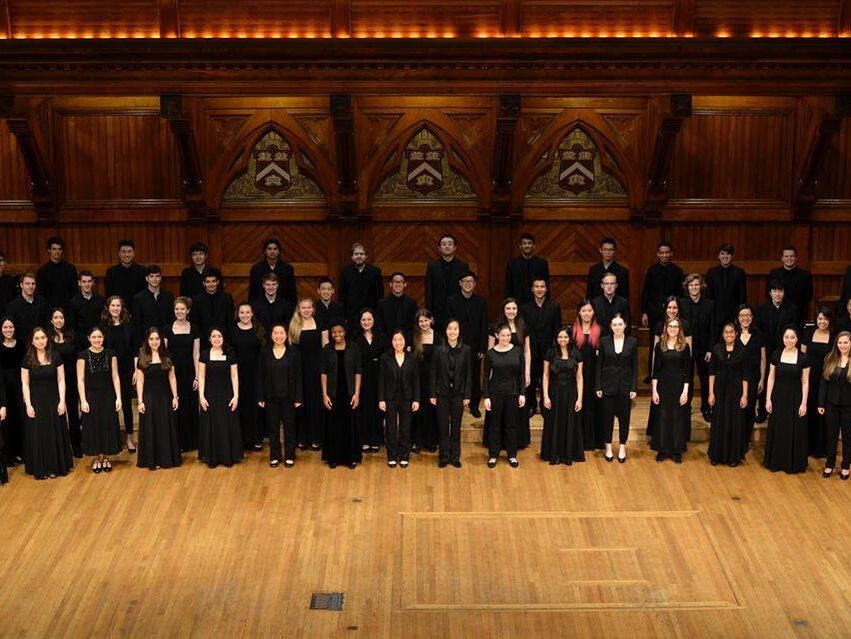 Harvard-Radcliffe Collegium Musicum singers standing on the Sanders Theatre stage in black concert attire. 