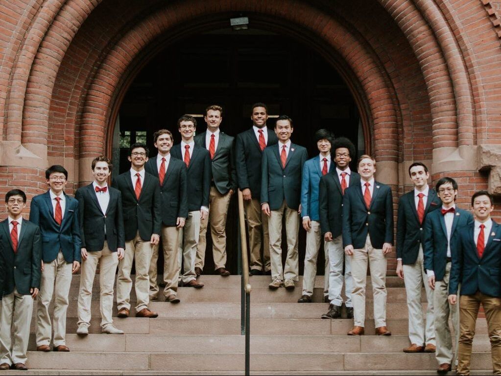 Harvard Glee Club Lite singers standing in an arch on steps in Harvard Yard, all wearing khaki pants, red ties, and navy blue blazers.