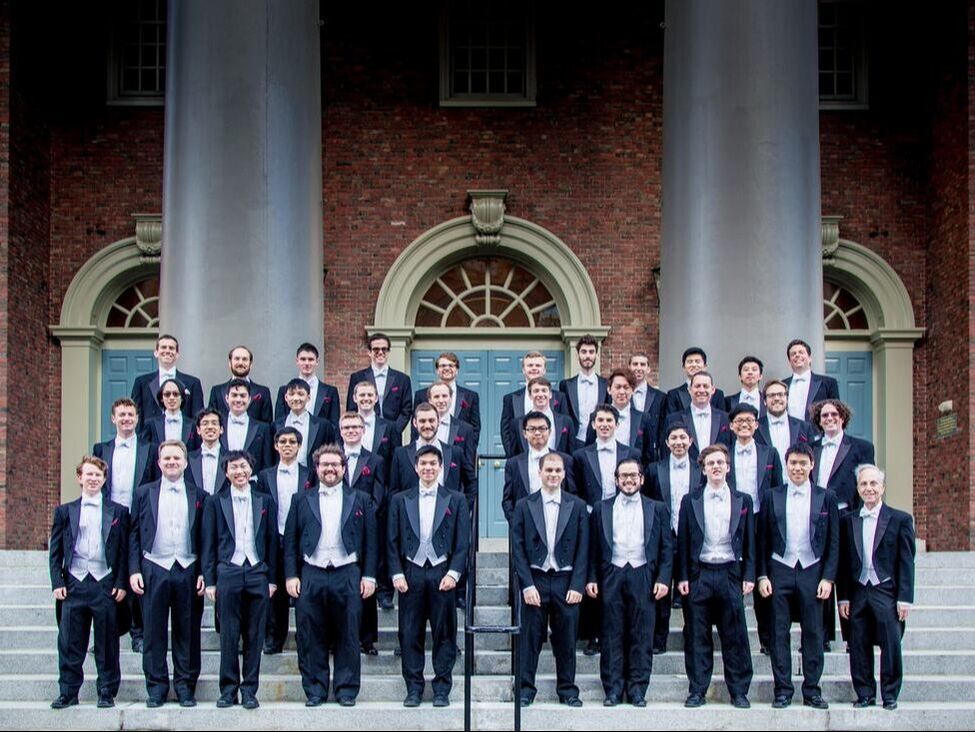Harvard Glee Club singers standing on the steps of Harvard Memorial Church in four rows. All are wearing tailcoat suits.