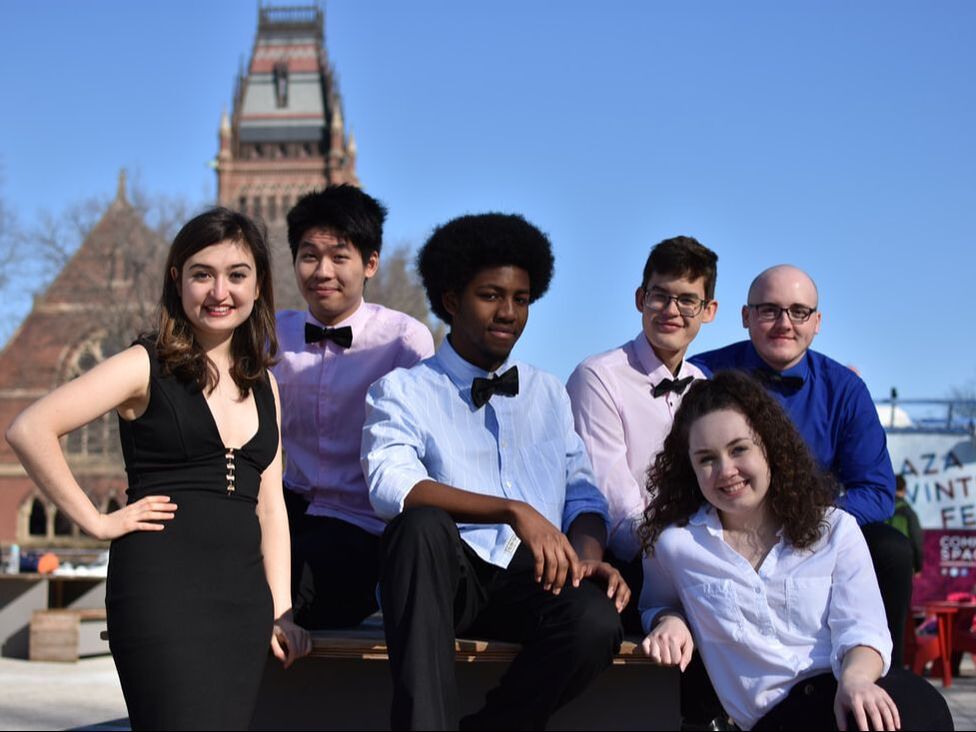 6 Collegium chamber singers sitting in Harvard Science Center Plaza with Memorial Hall in background.